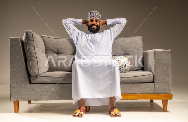 المباريات والألعاب الرياضية، الفرح لفوز الفريق المفضل، قضاء وقت ممتع بمتابعة المباراة، بورتريه لرجل عربي خليجي عماني يرتدي الدشداشة والكمة يجلس على أريكة مريحة معه كرة القدم ينظر إلى الكاميرا يضع يديه على رأسه بايماءات السرور والسعادة، خلفية بيج