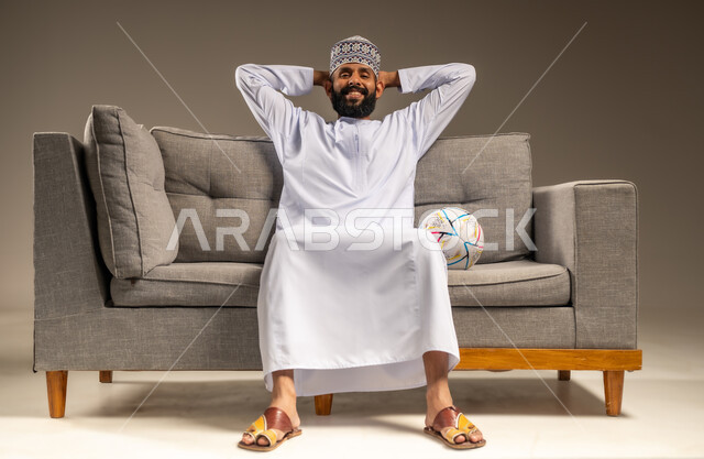 الفرح لفوز الفريق المفضل، قضاء وقت ممتع بمتابعة المباراة، بورتريه لرجل عربي خليجي عماني يرتدي الدشداشة والكمة يجلس على أريكة مريحة معه كرة القدم ينظر إلى الكاميرا يضع يديه على رأسه بايماءات السرور والسعادة، المباريات والألعاب الرياضية، خلفية بيج