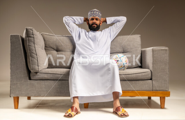 مفهوم الاستياء لعدم فوز الفريق المفضل، قضاء وقت ممتع بمتابعة المباراة، بورتريه لرجل عربي خليجي عماني يرتدي الدشداشة والكمة يجلس على أريكة مريحة معه كرة القدم ينظر إلى الكاميرا يضع يديه على رأسه بإيماءات الحزن والخسارة، المباريات والألعاب الرياضية، خلفية بيج