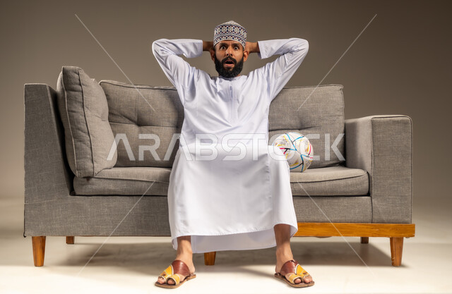 قضاء وقت ممتع بمتابعة المباراة، بورتريه لرجل عربي خليجي عماني يرتدي الدشداشة والكمة يجلس على أريكة مريحة معه كرة القدم ينظر إلى الكاميرا يضع يديه على رأسه بايماءات الصدمة والاندهاش، المباريات والألعاب الرياضية، خلفية بيج