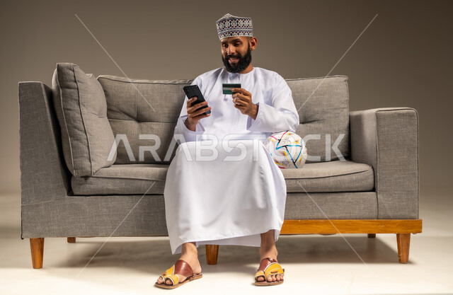 حجز التذاكر لمشاهدة الفريق المفضل عبر البطاقات الالكترونية، استخدام جهاز تقني حديث، قضاء وقت ممتع بمتابعة المباراة، بورتريه لرجل عربي خليجي عماني يرتدي الدشداشة والكمة يجلس على أريكة مريحة يمسك الهاتف المحمول والبطاقة البنكية، المباريات والألعاب الرياضية، خلفية بيج