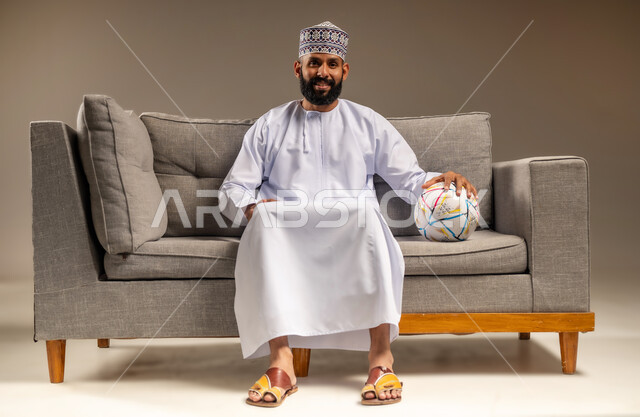 الاستمتاع بمشاهدة الفريق المفضل، قضاء وقت ممتع بمتابعة المباراة، بورتريه لرجل عربي خليجي عماني يرتدي الدشداشة والكمة يجلس على أريكة مريحة معه كرة القدم ينظر إلى الكاميرا بإيماءات السرور والثقة بالنفس، المباريات والألعاب الرياضية، خلفية بيج
