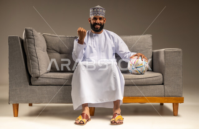 الاستمتاع بمشاهدة الفريق المفضل، قضاء وقت ممتع بمتابعة المباراة، بورتريه لرجل عربي خليجي عماني يرتدي الدشداشة والكمة يجلس على أريكة مريحة يمسك كرة القدم ينظر إلى الكاميرا بتعابير الحماس، رفع قبضة اليد بإيماءات الفوز والانتصار، المباريات والألعاب الرياضية، خلفية بيج