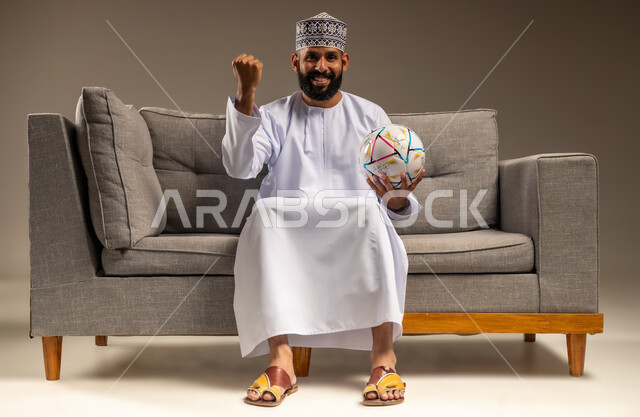 رفع قبضة اليد بإيماءات الفوز والانتصار، المباريات والألعاب الرياضية، الاستمتاع بمشاهدة الفريق المفضل، قضاء وقت ممتع بمتابعة المباراة، بورتريه لرجل عربي خليجي عماني يرتدي الدشداشة والكمة يجلس على أريكة مريحة يمسك كرة القدم ينظر إلى الكاميرا بتعابير الحماس، خلفية بيج