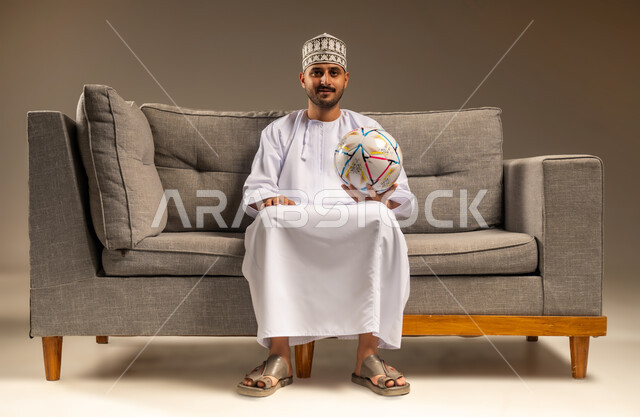 الاستمتاع بمشاهدة الفريق المفضل، قضاء وقت ممتع بمتابعة المباراة، بورتريه لرجل عربي خليجي عماني يرتدي الدشداشة والكمة يجلس على أريكة مريحة معه كرة القدم ينظر إلى الكاميرا بإيماءات السرور والثقة بالنفس، المباريات والألعاب الرياضية، خلفية بيج