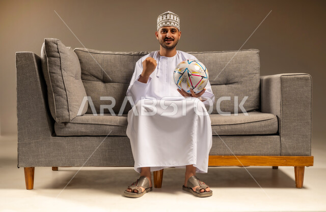 رفع قبضة اليد بإيماءات الفوز والانتصار، المباريات والألعاب الرياضية، الاستمتاع بمشاهدة الفريق المفضل، قضاء وقت ممتع بمتابعة المباراة، بورتريه لرجل عربي خليجي عماني يرتدي الدشداشة والكمة يجلس على أريكة مريحة يمسك كرة القدم ينظر إلى الكاميرا بتعابير الحماس، خلفية بيج