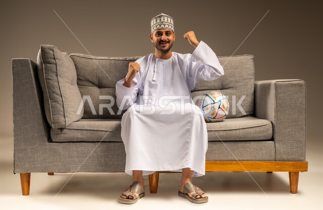 قضاء وقت ممتع بمتابعة المباراة، بورتريه لرجل عربي خليجي عماني يرتدي الدشداشة والكمة يجلس على أريكة مريحة معه كرة القدم ينظر إلى الكاميرا بتعابير الحماس، الاستمتاع بمشاهدة الفريق المفضل، رفع قبضتي اليدين بإيماءات الفوز والانتصار، المباريات والألعاب الرياضية، خلفية بيج