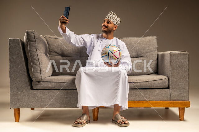 المباريات والألعاب الرياضية، التقاط الصور الذكارية، استخدام جهاز تقني حديث، قضاء وقت ممتع بمتابعة المباراة، بورتريه لرجل عربي خليجي عماني يرتدي الدشداشة والكمة يجلس على أريكة مريحة يمسك كرة القدم والهاتف المحمول يلتقط صورة سيلفي، خلفية بيج