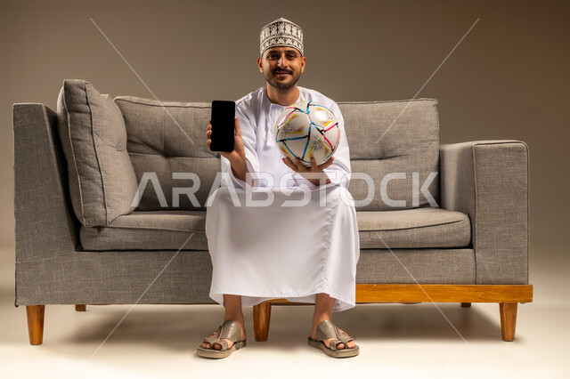 عرض شاشة سوداء فارغة، استخدام جهاز تقني حديث، قضاء وقت ممتع بمتابعة المباراة، بورتريه لرجل عربي خليجي عماني يرتدي الدشداشة والكمة يجلس على أريكة مريحة يمسك كرة القدم والهاتف المحمول ينظر إلى الكاميرا بإيماءات السرور، المباريات والألعاب الرياضية، خلفية بيج