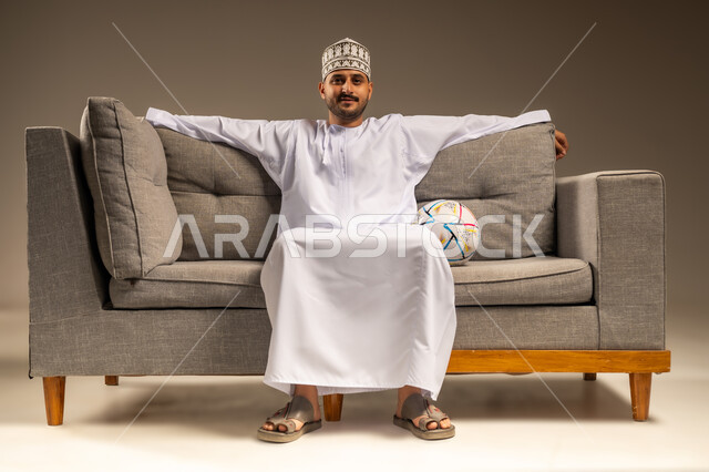 الاستمتاع بمشاهدة الفريق المفضل، قضاء وقت ممتع بمتابعة المباراة، بورتريه لرجل عربي خليجي عماني يرتدي الدشداشة والكمة يجلس على أريكة مريحة معه كرة القدم ينظر إلى الكاميرا بإيماءات السرور والثقة بالنفس، المباريات والألعاب الرياضية، خلفية بيج