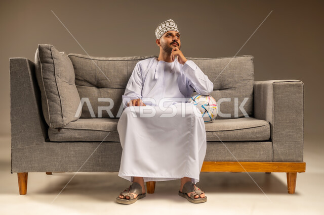 المباريات والألعاب الرياضية، قضاء وقت ممتع بمتابعة المباراة، بورتريه لرجل عربي خليجي عماني يرتدي الدشداشة والكمة يجلس على أريكة مريحة معه كرة القدم يضع يده أسفل ذقنه بايماءات التفكير والتخطيط، خلفية بيج