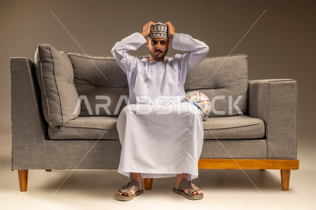 مفهوم الاستياء لعدم فوز الفريق المفضل، قضاء وقت ممتع بمتابعة المباراة، بورتريه لرجل عربي خليجي عماني يرتدي الدشداشة والكمة يجلس على أريكة مريحة معه كرة القدم ينظر إلى الكاميرا يضع يديه على رأسه بإيماءات الحزن والخسارة، المباريات والألعاب الرياضية، خلفية بيج