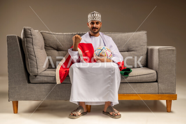 Raising a fist with a victory gesture, having a good time watching the match, portrait of an Arab Gulf Omani man wearing a dishdasha and a kummah sitting on a comfortable sofa with the flag of the Sultanate of Oman on his shoulders holding a football in his hand watching his favorite team, matches and sports games, beige background