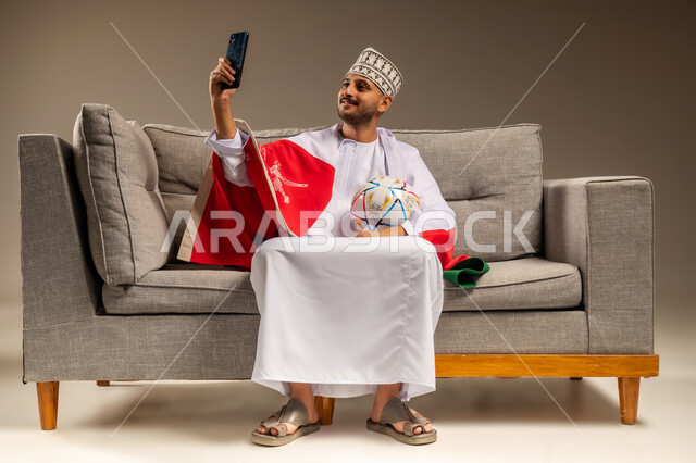 التقاط الصور الذكارية، استخدام جهاز تقني حديث، قضاء وقت ممتع بمتابعة المباراة، بورتريه لرجل عربي خليجي عماني يرتدي الدشداشة والكمة يجلس على أريكة مريحة يضع علم سلطنة عمان على كتفيه يمسك كرة القدم والهاتف المحمول يقوم بالتقاط صورة سيلفي، خلفية بيج