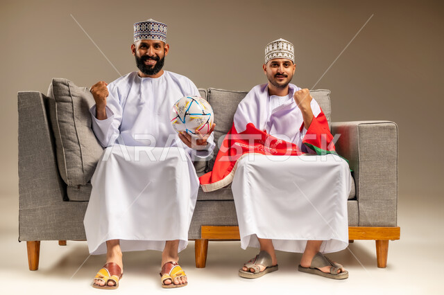 قضاء وقت ممتع بمتابعة المباراة مع الأصدقاء، بورتريه لرجلين عربيين عمانيين خليجيين يرتديان الدشداشة والكمة يجلسان على أريكة مريحة يضع أحدهما علم سلطنة عمان على كتفيه يقومان بمشاهدة الفريق المفضل بتعابير الاندماج، رفع قبضة اليد بإيماءات الفوز والانتصار، خلفية بيج