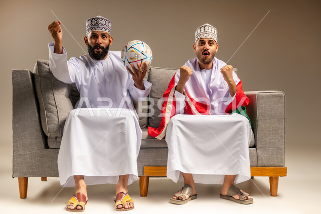 رفع قبضة اليد بإيماءات الفوز والانتصار، قضاء وقت ممتع بمتابعة المباراة مع الأصدقاء، بورتريه لرجلين عربيين عمانيين خليجيين يرتديان الدشداشة والكمة يجلسان على أريكة مريحة يضع أحدهما علم سلطنة عمان على كتفيه يقومان بمشاهدة الفريق المفضل بتعابير الاندماج، خلفية بيج