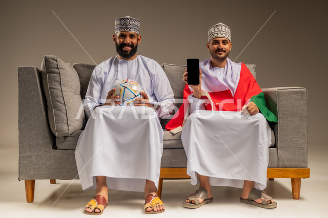 استخدام جهاز تقني حديث، عرض شاشة سوداء فارغة، قضاء وقت ممتع بمتابعة المباراة مع الأصدقاء، بورتريه لرجلين عربيين عمانيين خليجيين يرتديان الدشداشة والكمة يجلسان على أريكة مريحة يمسك أحدهما الهاتف المحمول ويضع علم سلطنة عمان على كتفيه، خلفية بيج
