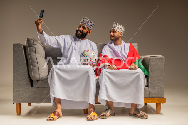 التقاط صور سيلفي تذكارية، استخدام جهاز تقني حديث، قضاء وقت ممتع بمتابعة المباراة مع الأصدقاء، بورتريه لرجلين عربيين عمانيين خليجيين يرتديان الدشداشة والكمة يجلسان على أريكة مريحة يمسك أحدهما الهاتف المحمول ويضع الآخر علم سلطنة عمان على كتفيه، خلفية بيج