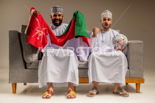 قضاء وقت ممتع بمتابعة المباراة مع الأصدقاء، بورتريه لرجلين عربيين عمانيين خليجيين يرتديان الدشداشة والكمة يجلسان على أريكة مريحة يمسك أحدهما علم سلطنة عمان يقومان بمشاهدة الفريق المفضل بتعابير الاندماج، رفع قبضة اليد بإيماءات الفوز والانتصار، خلفية بيج