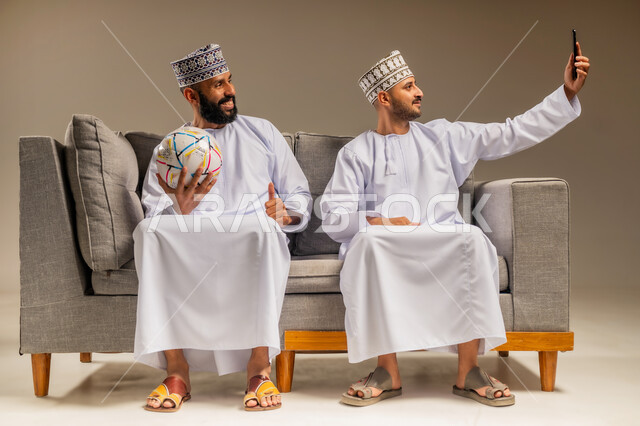 Using a modern technical device, portrait of two Arab Omani Gulf men wearing a dishdasha and a kummah sitting on a comfortable sofa, one of them holding a mobile phone taking a selfie with gestures of pleasure and happiness, taking souvenir photos, having a good time watching the match with friends, beige background