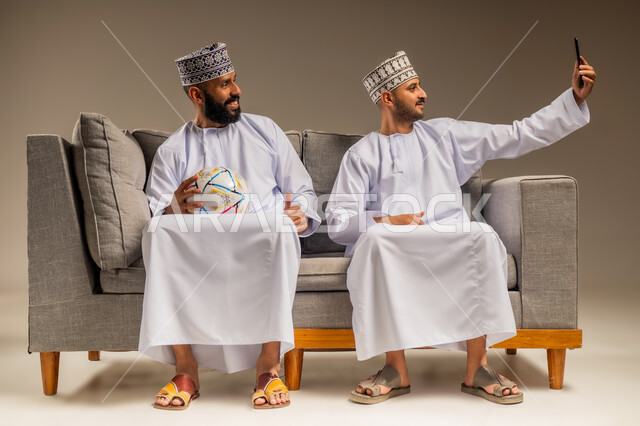 التقاط الصور التذكارية، قضاء وقت ممتع بمتابعة المباراة مع الأصدقاء، استخدام جهاز تقني حديث، بورتريه لرجلين عربيين عمانيين خليجيين يرتديان الدشداشة والكمة يجلسان على أريكة مريحة يمسك أحدهما هاتف محمول يلتقط صورة سيلفي بإيماءات السرور والسعادة، خلفية بيج