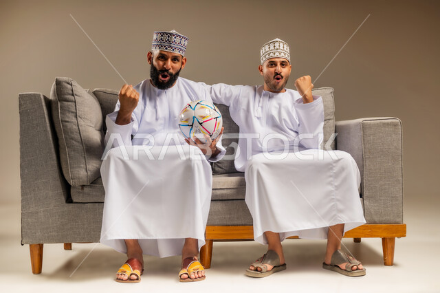 التفاعل والاندماج بمشاهدة الفريق المفضل, شاب يحمل كرة القدم، ايماءات الفرح والحماس بالانتصار, بورتريه لرجلين عربيين عمانيين خليجيين يرتديان الدشداشة والكمة يجلسان على أريكة مريحة يرفعان قبضة يدهما بتعابير الفوز, قضاء وقت ممتع بمتابعة المباراة, خلفية بيج