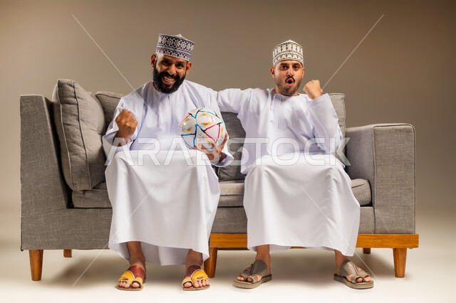 Gestures of joy and enthusiasm for victory, portrait of two Omani Gulf Arab men wearing dishdasha and kummah sitting on a comfortable sofa raising their fists with expressions of victory, interaction and integration while watching the favorite team, young man holding a football, having a good time watching the match, beige background