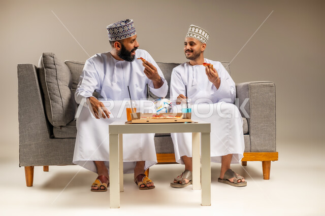 تبادل الحديث والحوار أثناء الطعام، مفهوم التسلية والترفيه، تناول وجبة سريعة ومشروب منعش, بورتريه لرجلين عربيين خليجيين عمانيين يرتديان الدشداشة والكمة يجلسان على أريكة مريحة يقومان بأكل البيتزا وشرب العصير، قضاء وقت ممتع مع الأصدقاء، خلفية بيج