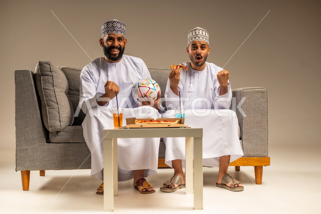 رفع قبضة اليد بإيماءات الفوز، قضاء وقت ممتع بمتابعة المباراة مع الأصدقاء, تناول وجبة سريعة ومشروب منعش، بورتريه لرجلين عربيين عمانيين خليجيين يرتديان الدشداشة والكمة يجلسان على أريكة مريحة يقومان بمشاهدة الفريق المفضل بتعابير الاندماج يمسك أحدهما كرة القدم، خلفية بيج