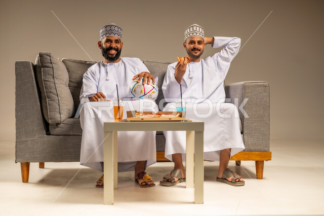 تناول وجبة سريعة ومشروب منعش، بورتريه لرجلين عربيين عمانيين خليجيين يرتديان الدشداشة والكمة يجلسان على أريكة مريحة يقومان بمشاهدة الفريق المفضل بتعابير التفاعل والاندماج يمسك أحدهما كرة القدم، قضاء وقت ممتع بمتابعة المباراة مع الأصدقاء, خلفية بيج