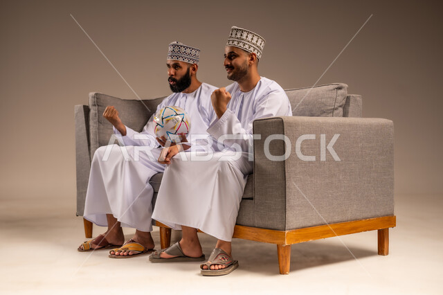 Gestures of joy and enthusiasm for victory, portrait of two Omani Gulf Arab men wearing dishdasha and kummah sitting on a comfortable sofa raising their fists with expressions of victory, interaction and integration while watching the favorite team, young man holding a football, having a good time watching the match, beige background