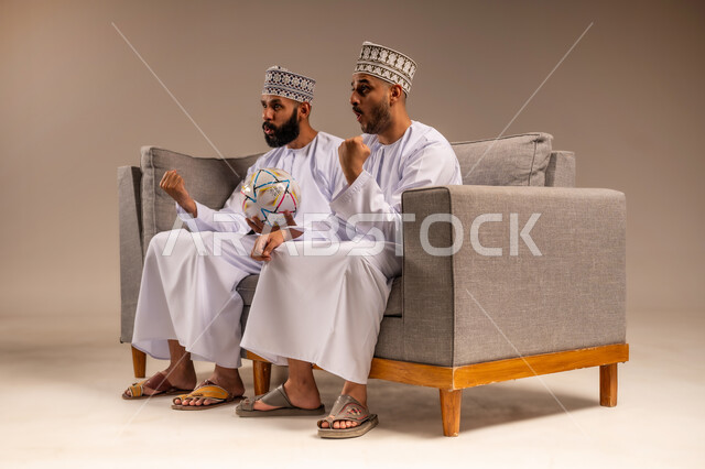 قضاء وقت ممتع بمتابعة المباراة, ايماءات الفرح والحماس بالانتصار, بورتريه لرجلين عربيين عمانيين خليجيين يرتديان الدشداشة والكمة يجلسان على أريكة مريحة يرفعان قبضة يدهما بتعابير الفوز, التفاعل والاندماج بمشاهدة الفريق المفضل, شاب يحمل كرة القدم، خلفية بيج