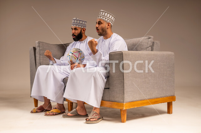 التفاعل والاندماج بمشاهدة الفريق المفضل, ايماءات الفرح والحماس بالانتصار, بورتريه لرجلين عربيين عمانيين خليجيين يرتديان الدشداشة والكمة يجلسان على أريكة مريحة يرفعان قبضة يدهما بتعابير الفوز, شاب يحمل كرة القدم، قضاء وقت ممتع بمتابعة المباراة, خلفية بيج