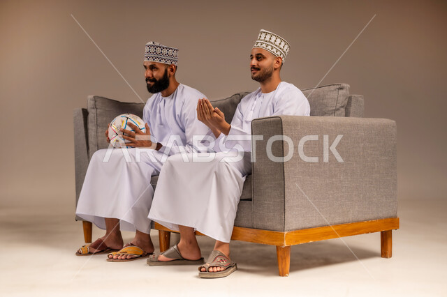 قضاء وقت ممتع بمتابعة المباراة مع الأصدقاء، إيماءات الدعاء والتضرع، المباريات والألعاب الرياضية، بورتريه لرجلين عربيين عمانيين خليجيين يرتديان الدشداشة والكمة يجلسان على أريكة مريحة يمسك أحدهما كرة القدم يقومان بمشاهدة الفريق المفضل بتعابير الاندماج، خلفية بيج