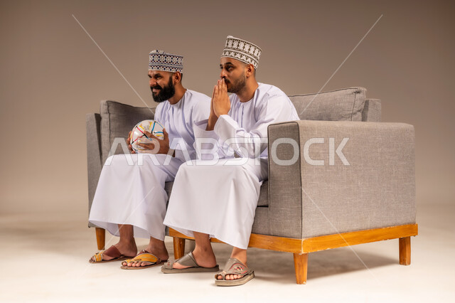 إيماءات الترقب وانتظار النتيجة، المباريات والألعاب الرياضية، قضاء وقت ممتع بمتابعة المباراة مع الأصدقاء، بورتريه لرجلين عربيين عمانيين خليجيين يرتديان الدشداشة والكمة يجلسان على أريكة مريحة يمسك أحدهما كرة القدم يقومان بمشاهدة الفريق المفضل بتعابير الاندماج، خلفية بيج