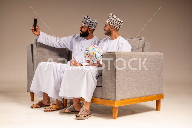 التقاط الصور التذكارية، استخدام جهاز تقني حديث، بورتريه لرجلين عربيين عمانيين خليجيين يرتديان الدشداشة والكمة يجلسان على أريكة مريحة يمسك أحدهما هاتف محمول يلتقط صورة سيلفي بإيماءات السرور والسعادة، قضاء وقت ممتع بمتابعة المباراة مع الأصدقاء، خلفية بيج