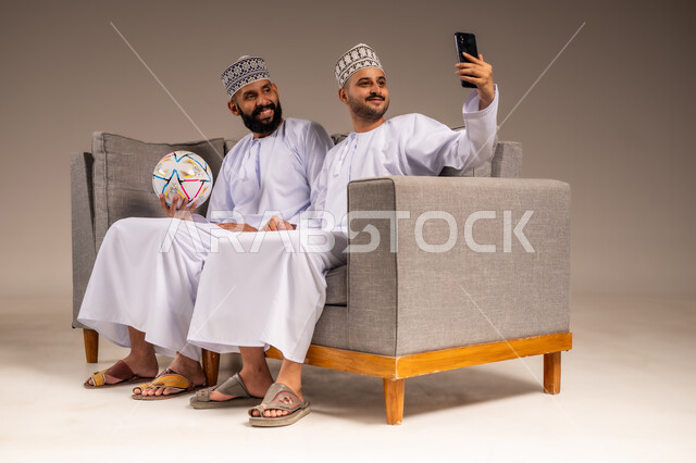 استخدام جهاز تقني حديث، بورتريه لرجلين عربيين عمانيين خليجيين يرتديان الدشداشة والكمة يجلسان على أريكة مريحة يمسك أحدهما هاتف محمول يلتقط صورة سيلفي بإيماءات السرور والسعادة، التقاط الصور التذكارية، قضاء وقت ممتع بمتابعة المباراة مع الأصدقاء، خلفية بيج