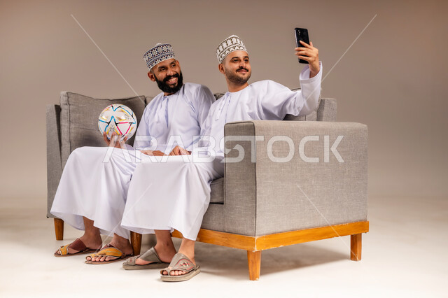 التقاط الصور التذكارية، قضاء وقت ممتع بمتابعة المباراة مع الأصدقاء، استخدام جهاز تقني حديث، بورتريه لرجلين عربيين عمانيين خليجيين يرتديان الدشداشة والكمة يجلسان على أريكة مريحة يمسك أحدهما هاتف محمول يلتقط صورة سيلفي بإيماءات السرور والسعادة، خلفية بيج