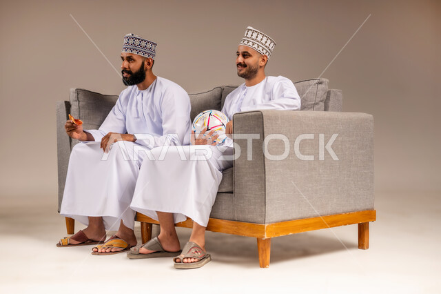 قضاء وقت ممتع بمتابعة المباراة مع الأصدقاء, تناول وجبة سريعة وسناك خفيف، بورتريه لرجلين عربيين عمانيين خليجيين يرتديان الدشداشة والكمة يجلسان على أريكة مريحة يقومان بمشاهدة الفريق المفضل بتعابير الاندماج يمسك أحدهما كرة القدم، المباريات والألعاب الرياضية، خلفية بيج