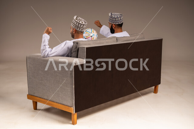 قضاء وقت ممتع بمتابعة المباراة, ايماءات الفرح والحماس بالانتصار, بورتريه من الجانب لرجلين عربيين عمانيين خليجيين يرتديان الدشداشة والكمة يجلسان على أريكة مريحة يرفعان قبضة يدهما بتعابير الفوز, التفاعل والاندماج بمشاهدة الفريق المفضل, شاب يحمل كرة القدم، خلفية بيج