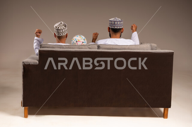 التفاعل والاندماج بمشاهدة الفريق المفضل, ايماءات الفرح والحماس بالانتصار, بورتريه من الخلف لرجلين عربيين عمانيين خليجيين يرتديان الدشداشة والكمة يجلسان على أريكة مريحة يرفعان قبضة يدهما بتعابير الفوز, شاب يحمل كرة القدم، قضاء وقت ممتع بمتابعة المباراة, خلفية بيج