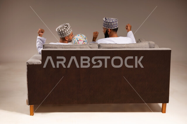 Interaction and integration while watching the favorite team, gestures of joy and enthusiasm for victory, portrait from the back of two Omani Gulf Arab men wearing dishdasha and kummah sitting on a comfortable sofa raising their fists with expressions of victory, young man holding a football, having a good time watching the match, beige background