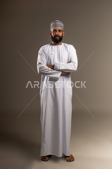 مفهوم الأناقة والرجولة، بورتريه لرجل عربي خليجي عماني يرتدي الدشداشة والكمة يقف بأيدي متقاطعة ينظر إلى الكاميرا بإيماءات الجدية والثقة بالنفس، الاهتمام بالمظهر الخارجي، الجسم بالطول الكامل، خلفية بيج