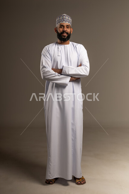 Concern for appearance, concept of elegance and masculinity, portrait of an Arab Gulf Omani man wearing a dishdasha and a kummah standing with crossed hands looking at the camera with gestures of pleasure and self-confidence, full-length body, beige background