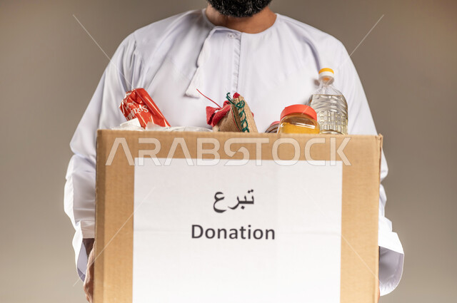 التقرب إلى الله بالعطاء والصدقة للمحتاجين، مفهوم المساعدة والتبرع لمشروع السلة الخيرية، بورتريه مقرب لرجل عربي خليجي عماني يرتدي الدشداشة يحمل صندوق من المواد الغذائية، جمعية ومبادرة إنسانية في سلطنة عمان، توفير الطعام للفقراء، خلفية بيج