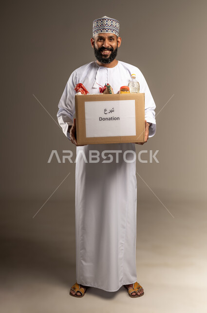 The concept of helping and donating to the charity basket project, a portrait of an Arab Gulf Omani man wearing a dishdasha and a kummah, holding a box of food supplies, a humanitarian association and initiative in the Sultanate of Oman, providing food to the poor, getting closer to God by giving and charity to the needy, full-length body, beige background