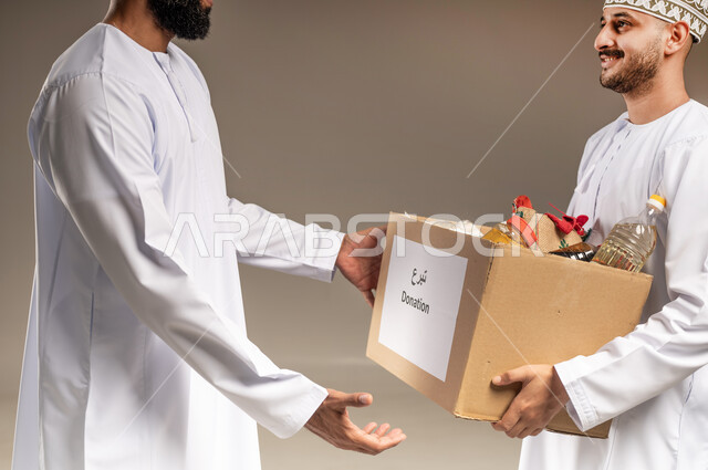 التقرب إلى الله بالعطاء للمحتاجين، المساعدة والتبرع لمشروع السلة الخيرية، بورتريه مقرب لرجل عربي خليجي عماني يرتدي الدشداشة يقوم بتقديم صندوق من المواد الغذائية لأحد المتعففين، مبادرة إنسانية في سلطنة عمان، توفير الطعام للفقراء، خلفية بيج