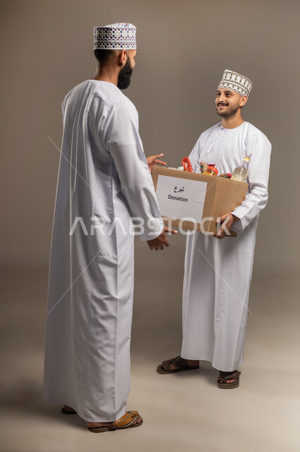 توفير الطعام للفقراء، التقرب إلى الله بالعطاء للمحتاجين، المساعدة والتبرع لمشروع السلة الخيرية، بورتريه لرجل عربي خليجي عماني يرتدي الدشداشة والكمة يقوم بتقديم صندوق من المواد الغذائية لأحد المتعففين، مبادرة إنسانية في سلطنة عمان، الجسم بالطول الكامل، خلفية بيج