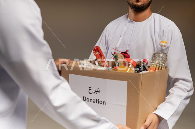 مبادرة إنسانية في سلطنة عمان، توفير الطعام للفقراء، التقرب إلى الله بالعطاء للمحتاجين، المساعدة والتبرع لمشروع السلة الخيرية، بورتريه مقرب لرجل عربي خليجي عماني يرتدي الدشداشة يقوم بتقديم صندوق من المواد الغذائية لأحد المتعففين، خلفية بيج