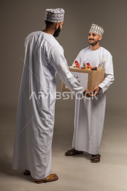 مبادرة إنسانية في سلطنة عمان، توفير الطعام للفقراء، التقرب إلى الله بالعطاء للمحتاجين، المساعدة والتبرع لمشروع السلة الخيرية، بورتريه لرجل عربي خليجي عماني يرتدي الدشداشة والكمة يقوم بتقديم صندوق من المواد الغذائية لأحد المتعففين، الجسم بالطول الكامل، خلفية بيج